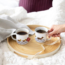 Load image into Gallery viewer, Two enamel mugs on a tray. The tray is placed on a bed and a hand is poised ready to pick up one of the mugs.