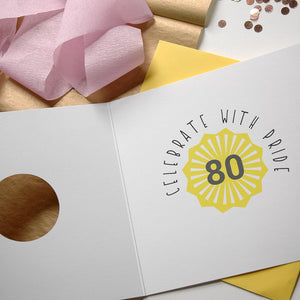 Greeting card with 'Celebrate with Pride 80' design on a wooden surface with pink flowers and confetti.