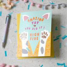 Load image into Gallery viewer, A greeting card with a playful cat-themed design reading 'AMAZING YAY YOU DID IT! HIGH FIVE' is photographed on a light marble-like surface with confetti, a pen, and pom-pom garland around it. The card shows two raised paw prints in white, grey, and brown on a pale blue background, with orange and pink text and yellow/orange lower sections.