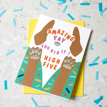 Load image into Gallery viewer, A colorful greeting card featuring a high-five dog design with the text 'AMAZING YAY YOU DID IT! HIGH FIVE', placed on top of a yellow envelope on a light marble surface with scattered confetti.