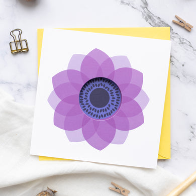 A greeting card with a purple anemone flower design in a circle cut format, accompanied by a yellow envelope, displayed on a marble surface with clips and a pen.