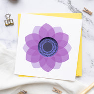 A greeting card with a purple anemone flower design in a circle cut format, accompanied by a yellow envelope, displayed on a marble surface with clips and a pen.
