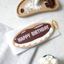 Load image into Gallery viewer, Birthday card shaped as an eclair with a bread bag envelope. On a marble surface surrounded by ingredients - icing sugar and chocolate on a wooden tray and whipped cream in a bowl.