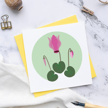 Load image into Gallery viewer, Small square greetings card with an Illustration of a cyclamen with a bud either side and 3 leaves.  This is plaved on a marble surface with a yellow envelope and a pen nib.
