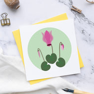Small square greetings card with an Illustration of a cyclamen with a bud either side and 3 leaves.  This is plaved on a marble surface with a yellow envelope and a pen nib.