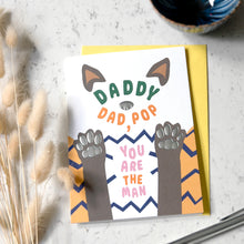 Load image into Gallery viewer, A greeting card with a humorous 'Daddy Dad, Pop' design featuring cartoon paws and the phrase 'You Are The Man', displayed on a yellow envelope over a light marble surface with dried bunny tail grass and a ceramic bowl.