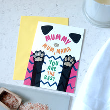Load image into Gallery viewer, A greeting card with a white background featuring a stylized dog face and paws in black, pink, blue, and green, with the text 'MUMMY MUM, MAMA YOU ARE THE BEST'. A yellow envelope is placed behind it on a light marble-like surface with a white candle partially visible in the background.