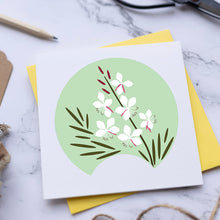Load image into Gallery viewer, A square white greeting card with a circular green illustration of white flowers and green leaves, placed on top of a yellow card on a marble surface with scissors, a pencil, and twine nearby.