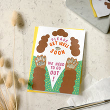 Load image into Gallery viewer, A square greeting card with a playful dog paw design, placed on a light marble surface with dried grass stems and a pen nearby. The card reads 'PLEASE GET WELL SOON' and 'WE NEED TO GO OUT!' with illustrated brown paws and green dotted borders.