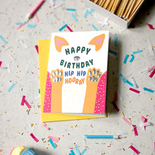 Load image into Gallery viewer, A colorful 'Happy Birthday Hip Hip Hoory' dog-themed greeting card with a yellow envelope, styled on a light grey surface scattered with confetti, with a box of matches in the background.