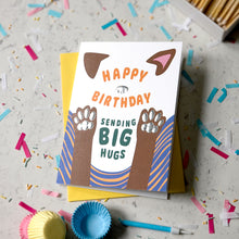 Load image into Gallery viewer, A dog-themed birthday card with a white front, featuring the text 'HAPPY BIRTHDAY SENDING BIG HUGS' and an illustration of dog paws and ears, displayed on top of a yellow envelope on a light grey confetti-covered surface.