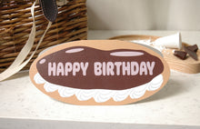Load image into Gallery viewer, A novelty birthday card shaped like a chocolate ceclair with the words 'HAPPY BIRTHDAY' in the shine of the chocolate, displayed on a light marble surface with a wicker basket and chocolate pieces in the background.