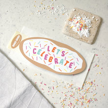 Load image into Gallery viewer, A decorative oval creetings card “LET’S CELEBRATE” with colorful sprinkle accents, shown on a bread bag envelope on a light stone surface. A small round “Baked Goods” brand seal appears on the left, and styled props including sprinkles, powdered sugar, and a linen cloth. 
