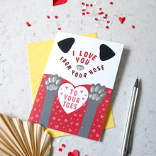 Load image into Gallery viewer, A novelty Valentine's Day card with a dog theme, featuring the text 'I LOVE YOU FROM YOUR NOSE TO YOUR TOES' and a cartoon dog face and paws. The card is displayed on a light gray marble surface with a yellow envelope, a pen, and scattered red heart confetti.