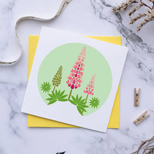 Load image into Gallery viewer, A square white art print featuring a circular botanical illustration of pink and green flowers, displayed on a yellow cardstock sheet over a white marble surface with decorative dried stems, a pen, and small wooden clothespins.