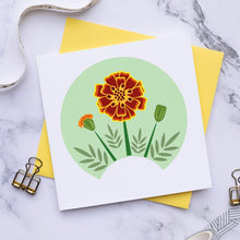 Load image into Gallery viewer, A square white art print card featuring a stylized orange and yellow marigold on a light green circular background, styled on a yellow card and marble surface with ribbon, clips, and washi tape.