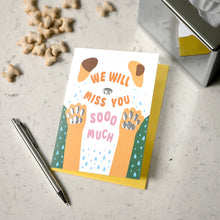 Load image into Gallery viewer, A dog-themed greeting card with the text 'WE WILL MISS YOU SOOO MUCH' and an illustration of paw prints, placed on a yellow backing card on a light speckled surface with a pen, small bone-shaped props, and a tissue box nearby.