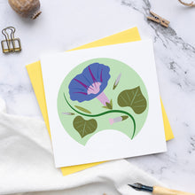 Load image into Gallery viewer, A square white greeting card featuring a stylized blue-purple morning glory flower with green leaves inside a pale green circle, layered over a yellow envelope. The card is arranged on a white marble surface with linen cloth, dried pods, binder clips, string, and a wooden pen nearby.