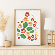 A framed botanical art print featuring red, orange, and green flowers with yellow centers, styled on a light wood shelf next to a white vase with dried grass and a woven table lamp.