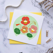 Load image into Gallery viewer, A square white greeting card featuring a stylized illustration of nasturtium flowers in red, orange, and pink with green leaves, layered over a yellow envelope on a white marble surface. A fountain pen, dried botanicals, and small wooden clothespins are arranged around it.