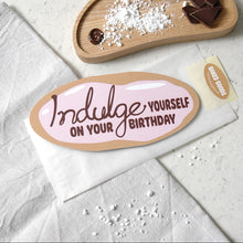 Load image into Gallery viewer, An oval birthday card with the phrase 'Indulge Yourself on Your Birthday' in brown  chocolate lettering on a pink icing background, placed on a bread bag envelope. A small sticker reading 'Baked Goods' is visible nearby, along with styled baking props including chocolate pieces, powdered sugar, and a wooden tray on a light marble surface.
