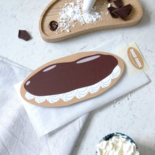 Load image into Gallery viewer, A flat, die-cut paper cutout of a chocolate cream puff pastry with white whipped cream detailing, displayed on a white napkin. The bread bag envelope features an oval sticker with a 'Baked Goods' brand tag.
