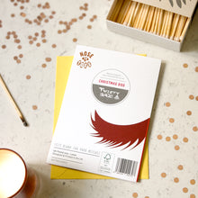Load image into Gallery viewer, A printed card labeled 'Christmas Dog' lies on top of a yellow envelope on a light speckled surface with scattered confetti. A matchbox with wooden matches and a lit candle are partially visible around it.