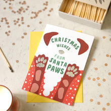 Load image into Gallery viewer, A Christmas-themed dog greeting card with the text 'CHRISTMAS WISHES FROM SANTA PAWS' and illustrations of red dog paws, placed on a yellow envelope over a light surface with confetti, a matchbox, and a candle visible around it.