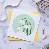 small card will illustration of 3 snowdrops. The card is on top of a yellow envelope on a white marble surface.