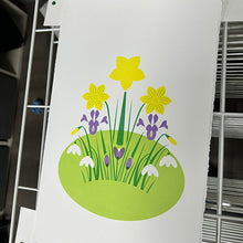 Load image into Gallery viewer, A white printed card or sign featuring a stylized spring flower illustration with yellow daffodils, purple irises, white snowdrops, green stems, and a light green oval ground, photographed against shelving.