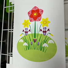 Load image into Gallery viewer, A white rectangular sign or print featuring a colorful spring flower illustration, including a large red-pink flower, yellow daisies, purple irises, white snowdrops, and green grass on a light green oval patch. The sign is photographed on a white wire rack.