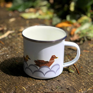 Enamel Mug on ground with mother duck facing towards the handle. The first baby duck is just visable.