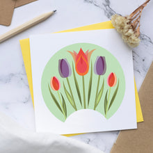 Load image into Gallery viewer, A square white greeting card with a colorful tulip illustration, layered over a yellow card and placed on a marble surface with kraft paper tags, a pencil, binder clips, and dried flowers as styled props.