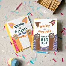 Load image into Gallery viewer, Two colorful birthday cards with dog paw illustrations are displayed on a light speckled surface with confetti, a matchbox, and small party props. One card says 'Happy Birthday Hip Hip Hooray!' and the other says 'Happy Birthday Sending Big Hugs'.