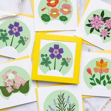 Load image into Gallery viewer, A styled flat-lay of multiple floral art cards or envelopes arranged on a light marble surface, with a white measuring tape draped around them. The cards feature circular botanical illustrations in pink, purple, orange, green, and yellow tones.